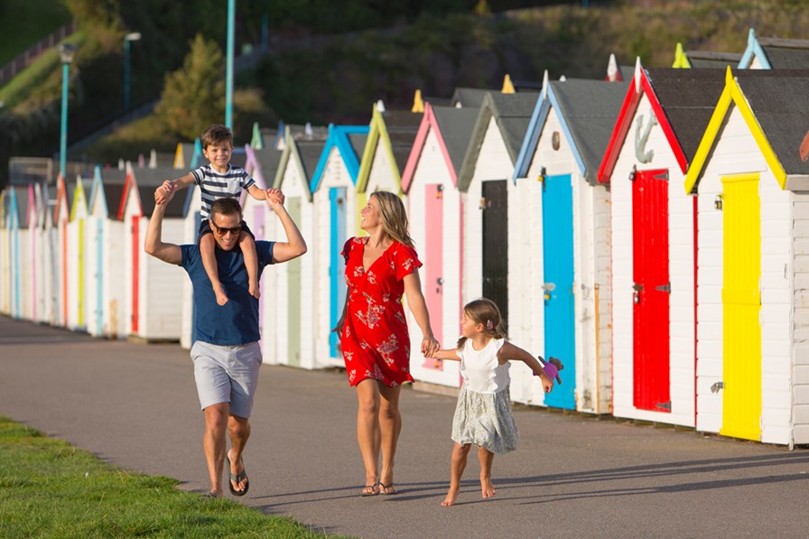 A family walking in front of beach huts. The father is carrying a young boy on his shoulders while the mother holds hand with a young girl.