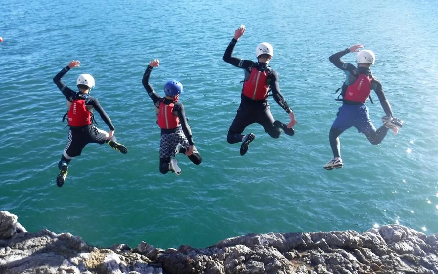 Four young people wearing wetsuits and safety helmets jumping of a cliff into the sea.