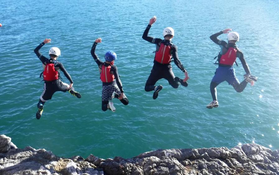 Four young people wearing wetsuits and safety helmets jumping of a cliff into the sea.